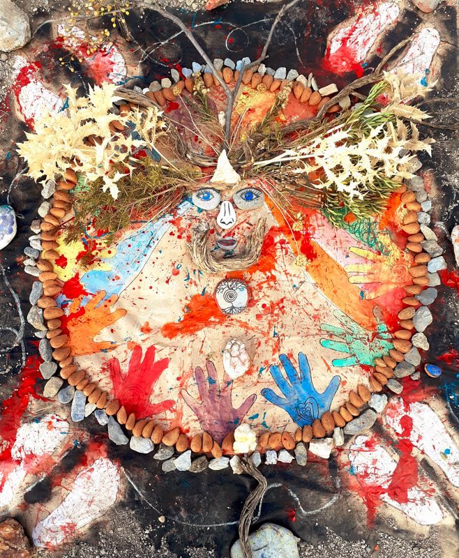 An outdoor bodymap edged in stones, with branches, dried flowers, and painted handprints